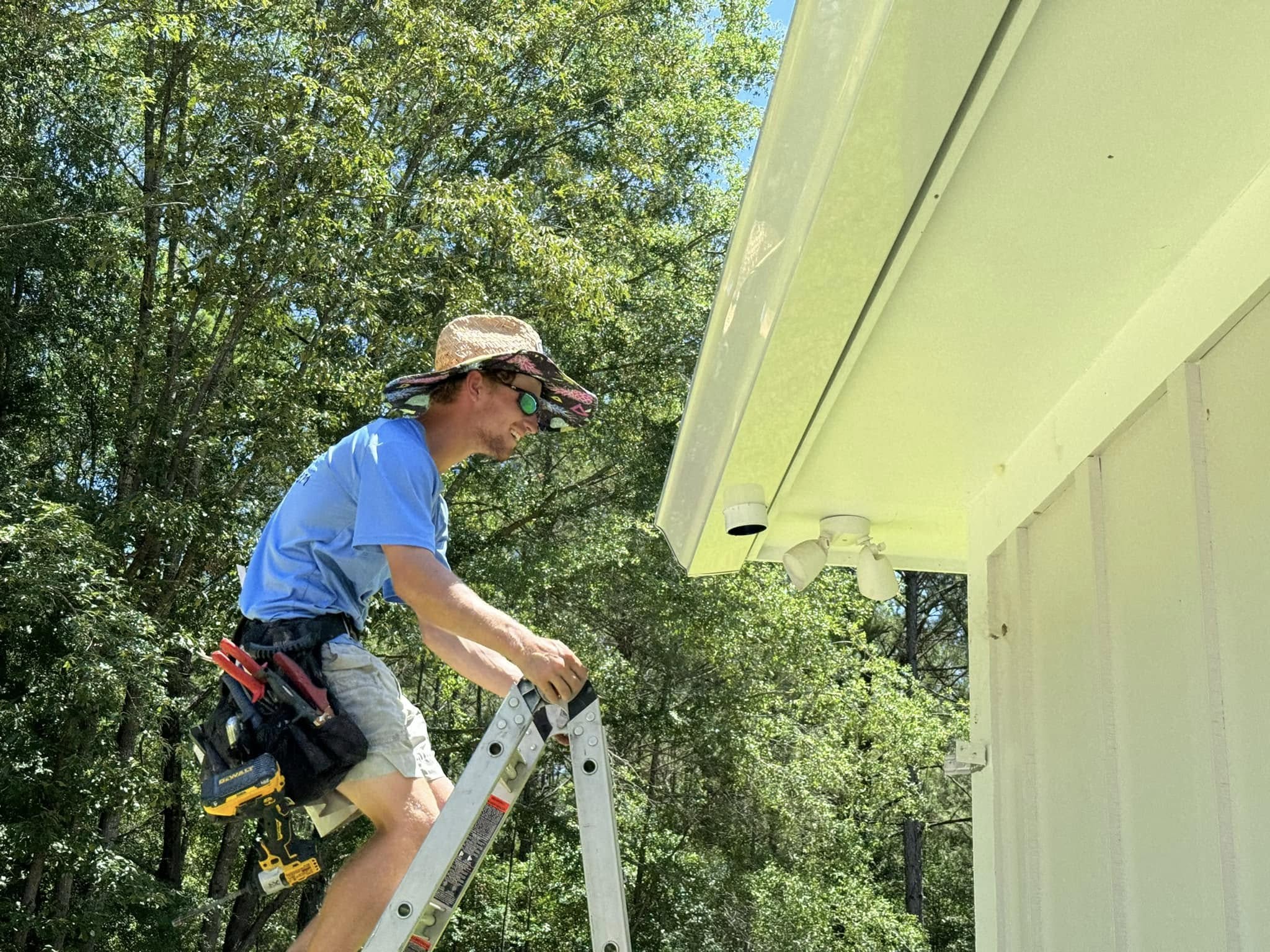 Worker on a ladder installing or repairing a gutter along the roofline of a house, with tools on a belt and trees in the background.