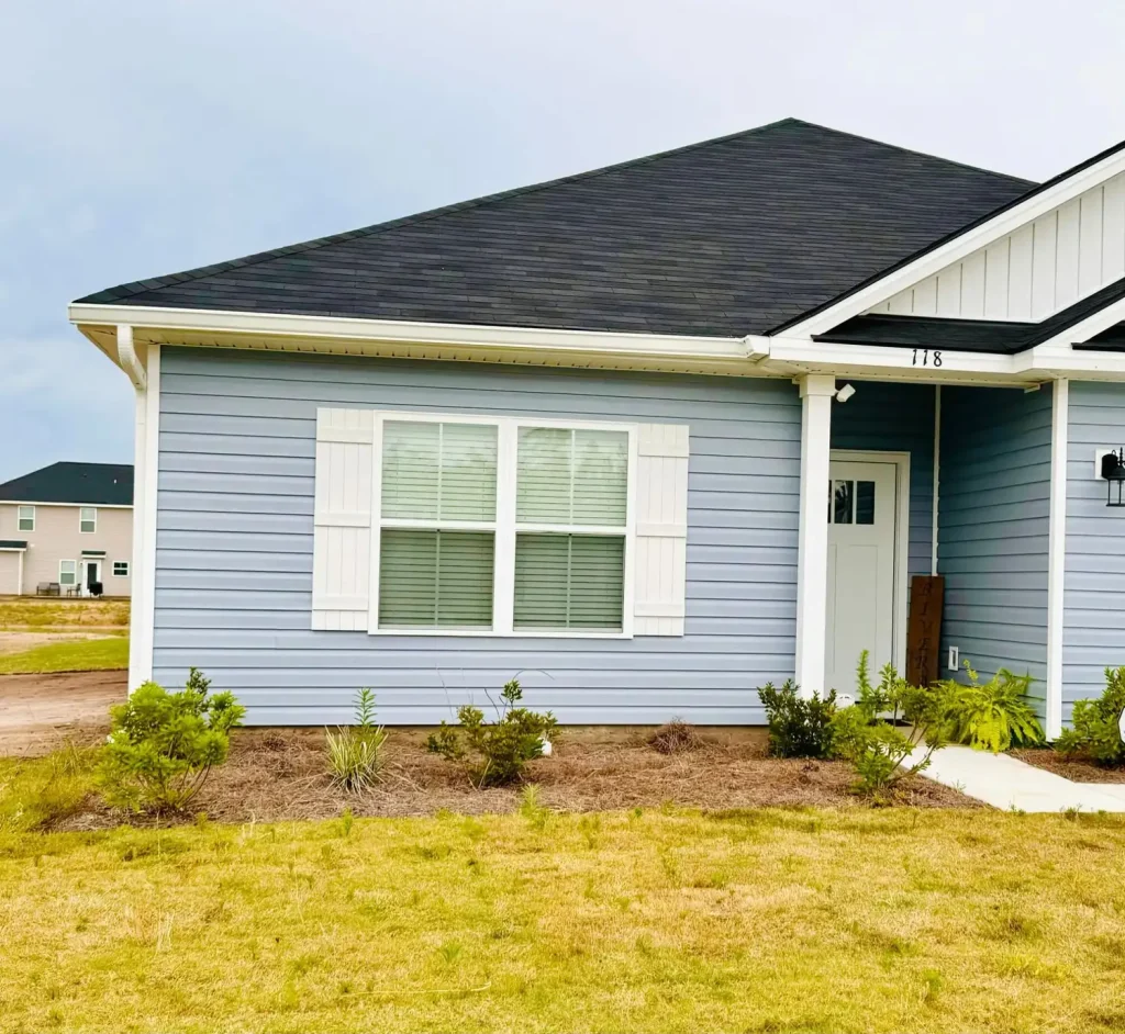 Single-story home with blue-gray siding, white shutters, a dark shingle roof, and white gutters, featuring a small front entry and landscaped yard.