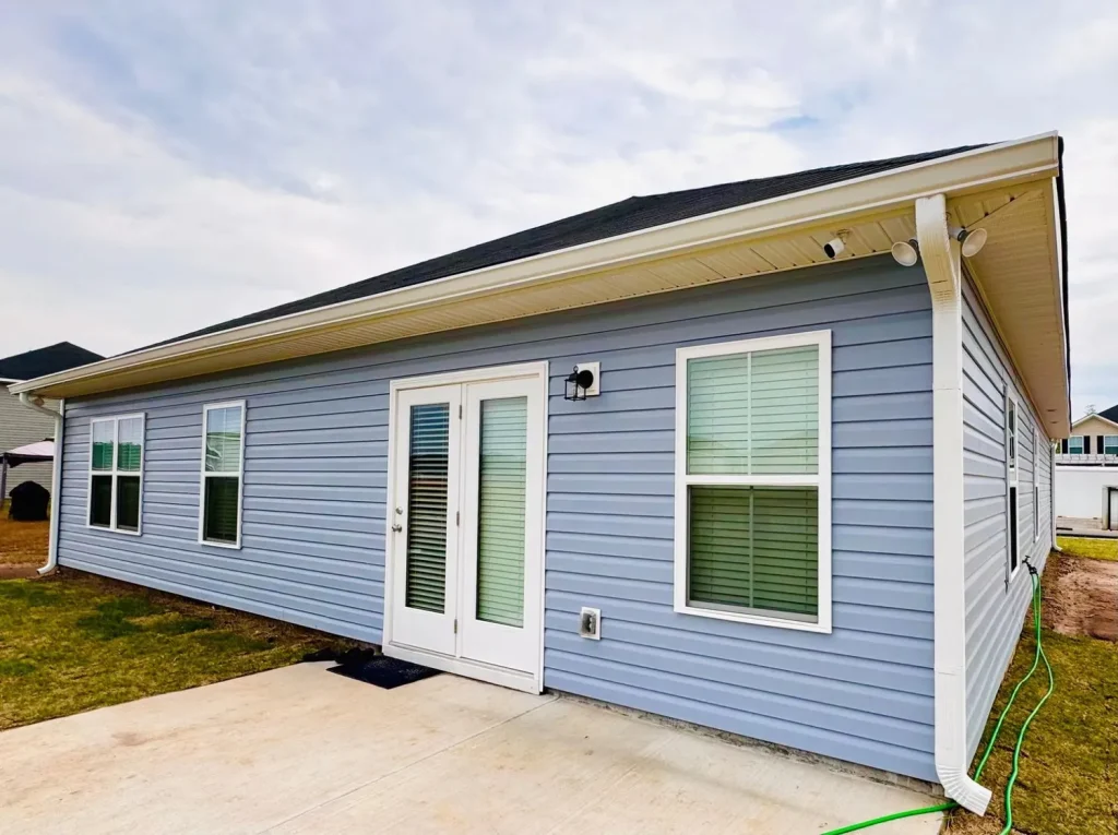 Rear view of a single-story blue house with white trim, featuring a sloped roof, gutters, double glass patio doors, and a small concrete patio under an overcast sky.