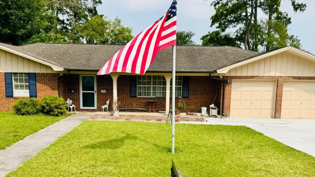 Single-story brick home with a shingle roof, white gutters, and a front porch, featuring a large American flag displayed in the front yard.
