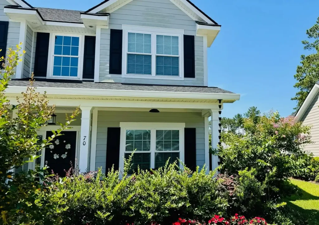 Two-story home with light siding, black shutters, a covered front porch, and white gutters, surrounded by dense landscaping and greenery.