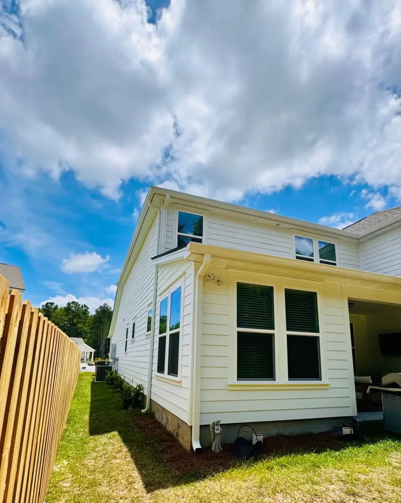 Two-story white house with horizontal siding, multiple windows, and a covered patio, viewed from the side yard with a wooden fence under a partly cloudy blue sky.