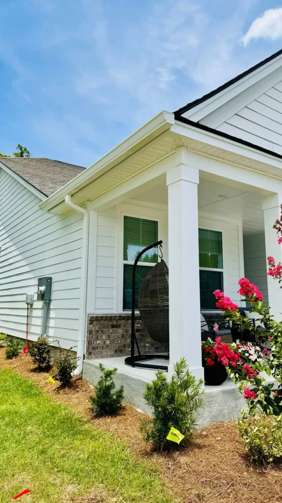 Front porch of a home with white siding, white gutters and downspout, and square columns, featuring a hanging chair and flowering plants nearby.