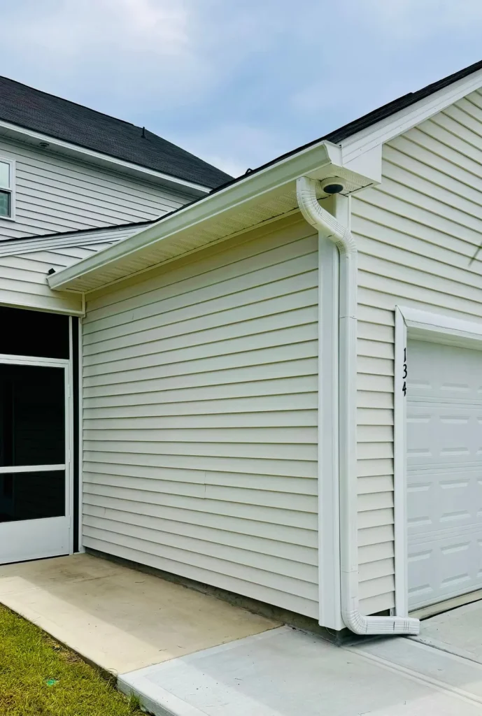 Close-up of a home’s exterior with light siding, featuring a white gutter and downspout running along the roofline next to a garage door.