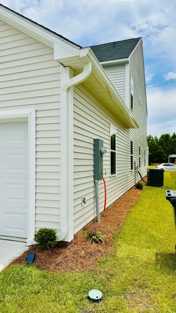 Side view of a home with light siding, white gutters, and utility connections on the exterior wall, set beside a grassy yard with an outdoor AC unit.
