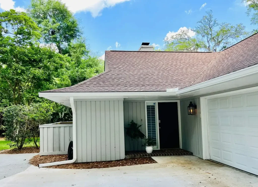 Single-story home with light vertical siding, a shingle roof, white gutters and downspout, and a garage, surrounded by trees and a driveway.