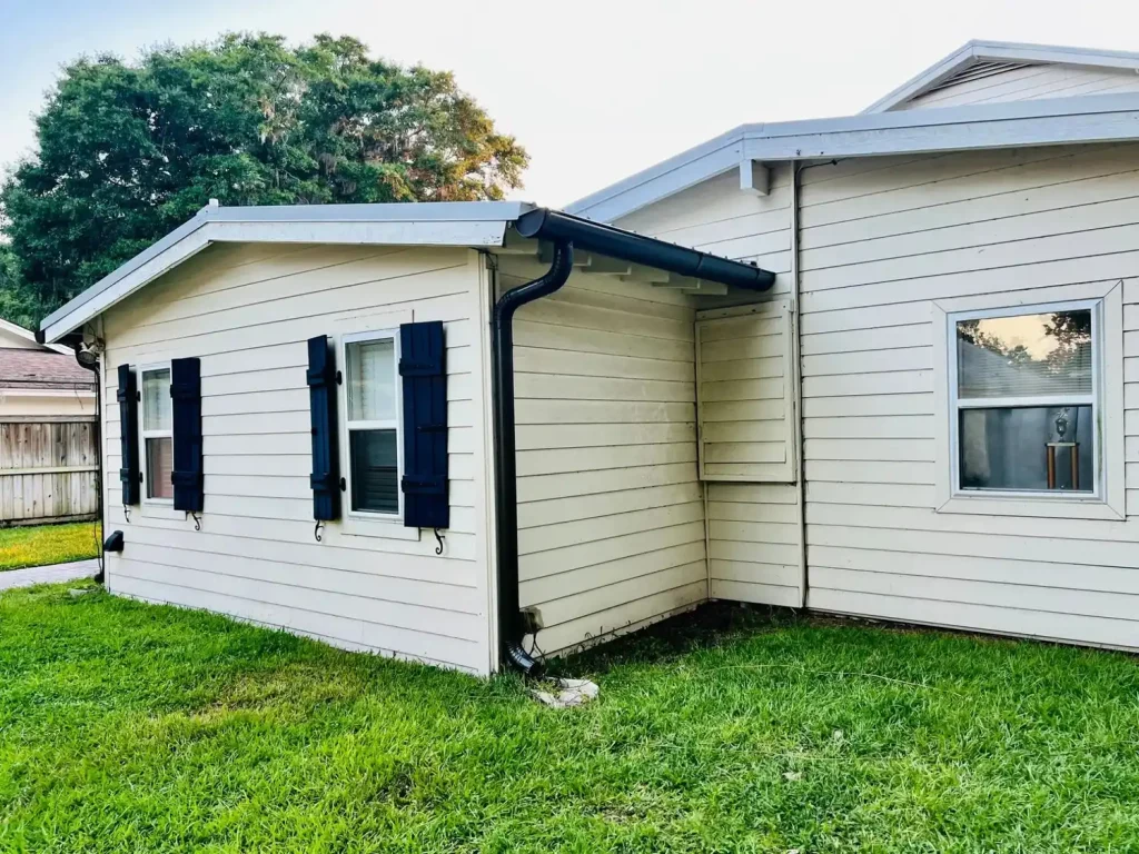 Single-story home with light siding, black shutters, and a black gutter and downspout system, set beside a grassy yard.
