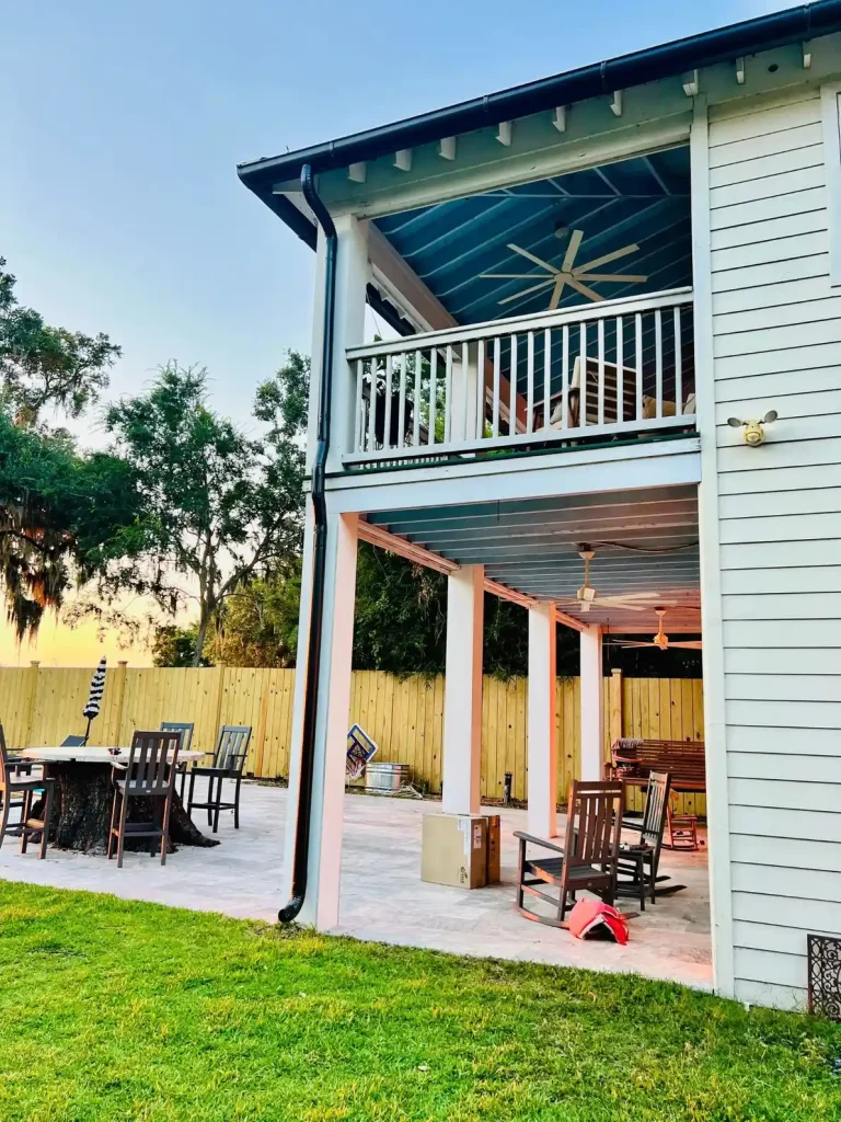 Two-story home with a covered balcony and patio below, featuring black gutters and downspouts, outdoor seating, and a fenced backyard.