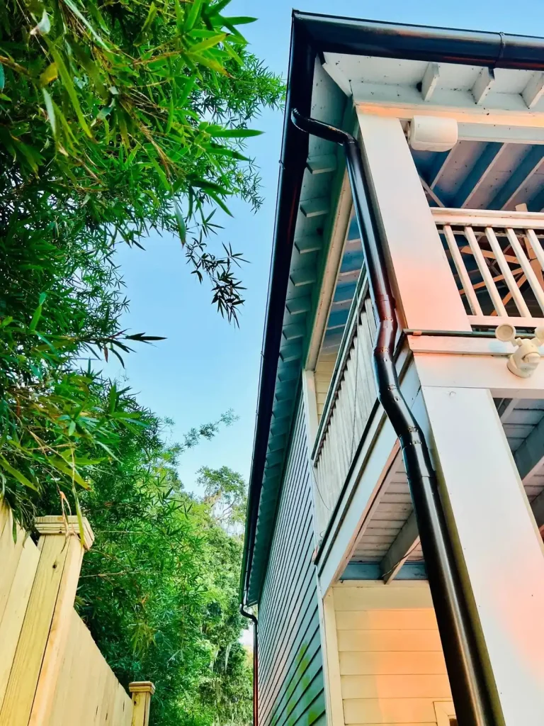 Upward view of a two-story home with a balcony and black gutter and downspout system, surrounded by trees and greenery.