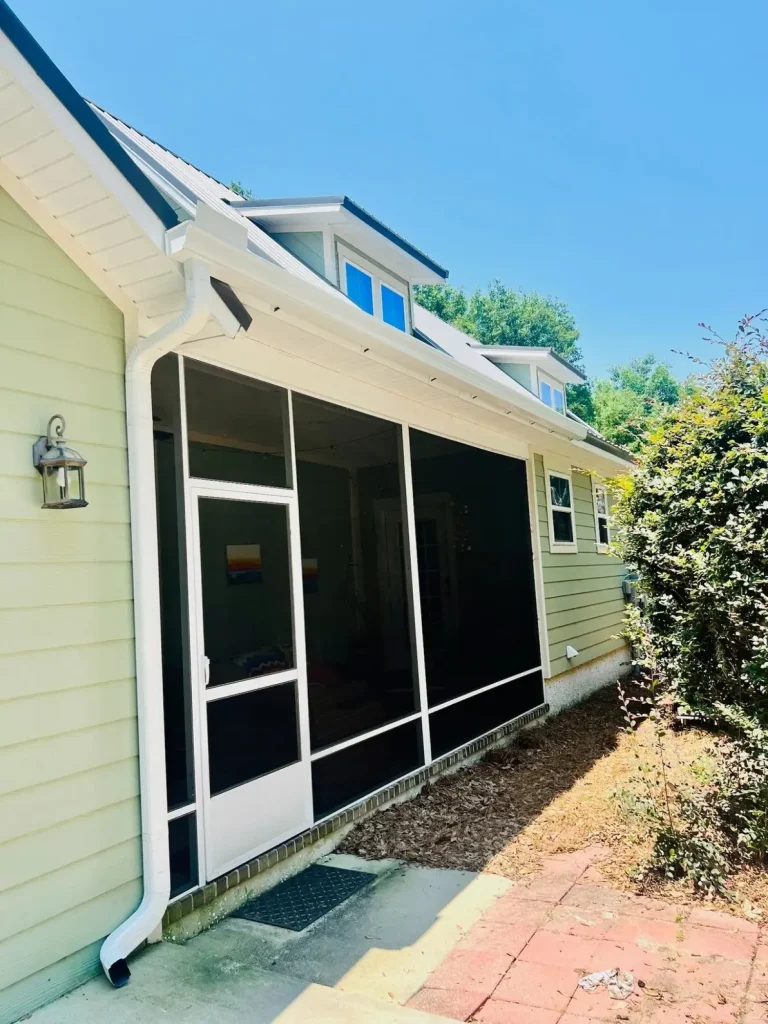 Screened-in porch on a light green home with white gutters and downspout, surrounded by shrubs and trees.