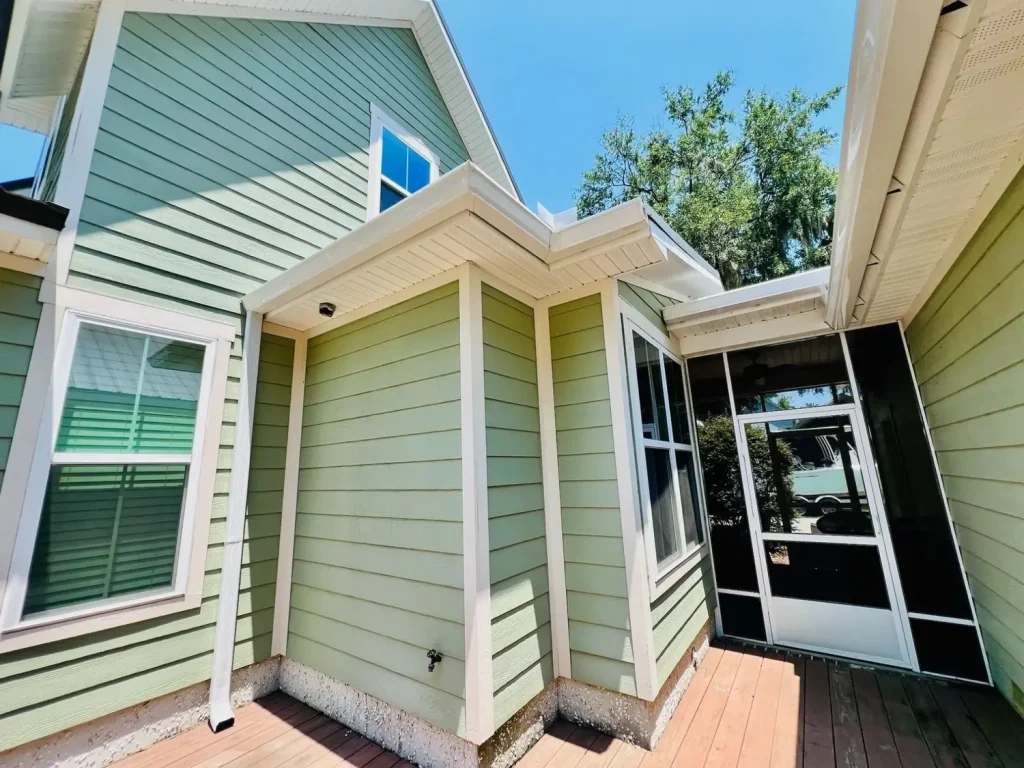 Close-up of a green two-story house exterior with white trim, showing rooflines, gutters, windows, and a screened porch entrance on a wooden deck under a clear blue sky.
