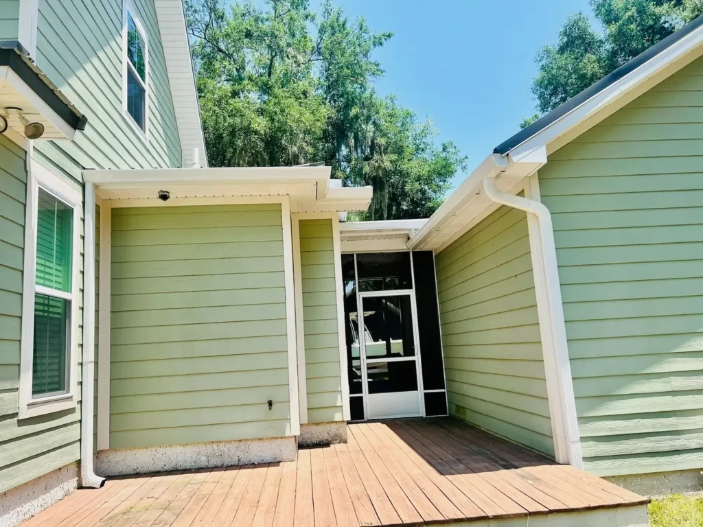 Back entry area of a home with light green siding, white gutters and downspouts, and a small wooden deck leading to a screened door, surrounded by trees.