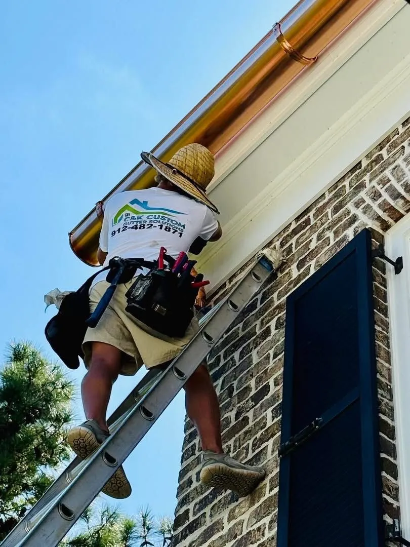 Worker on a ladder installing a copper gutter along the roofline of a brick house, wearing a tool belt and wide-brim hat.