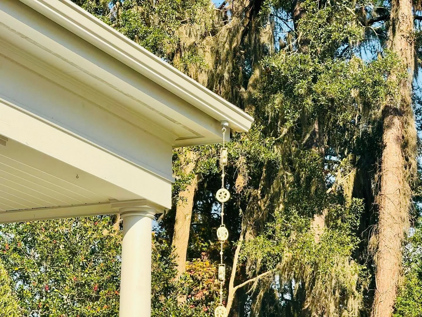 Corner of a house roof with white gutters and soffit, featuring a decorative rain chain hanging down, with trees in the background.