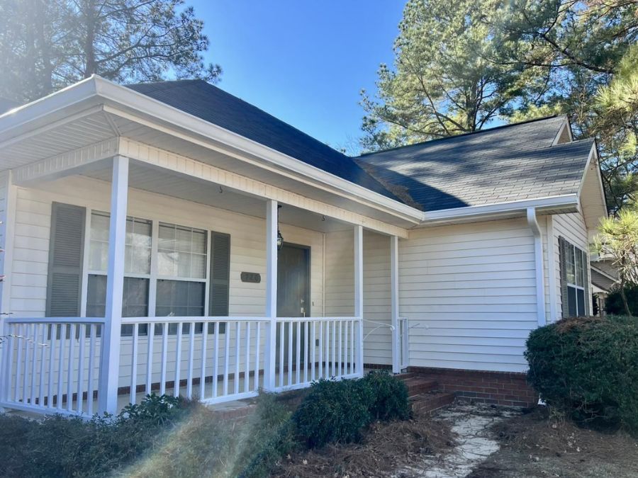 Small white house with a covered front porch, railing, and visible gutters along the roofline, surrounded by trees on a sunny day.