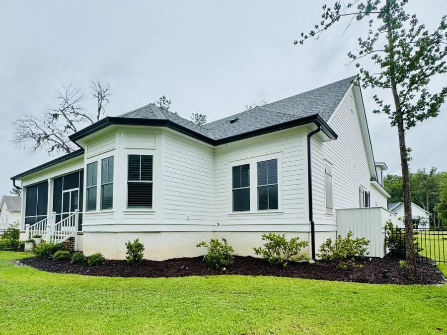 Single-story white home with black gutters and downspouts, a gray shingle roof, and landscaped yard with fresh mulch and small shrubs.