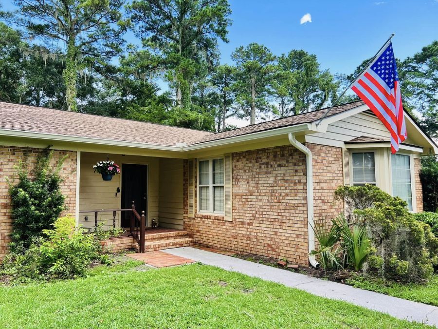 Single-story brick home with a shingle roof, white gutters, front porch entry, and an American flag mounted near the corner, surrounded by lawn and trees.