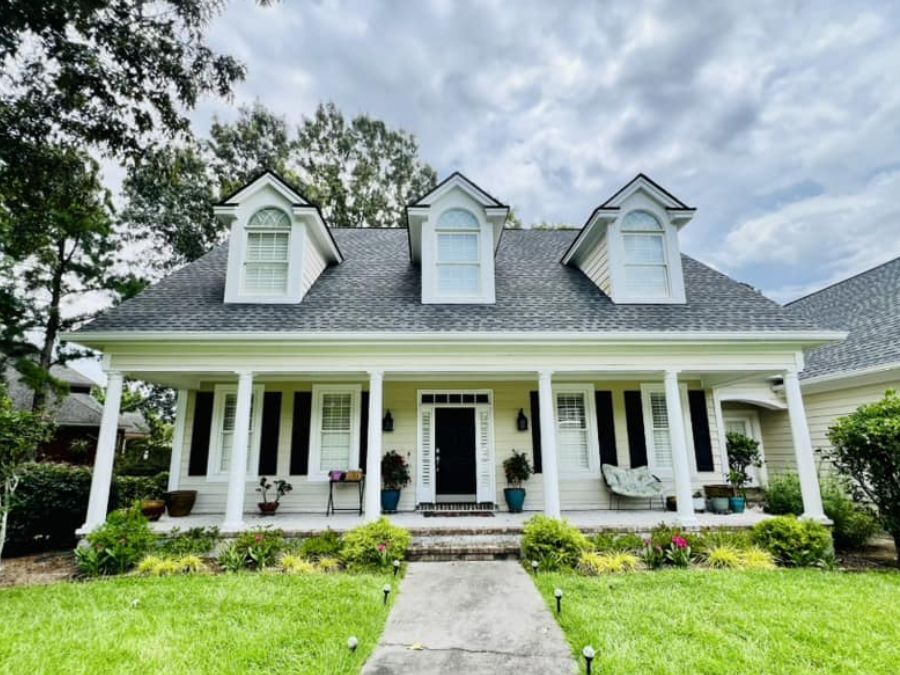 Front view of a traditional white house with a large covered porch, columns, dormer windows, and a landscaped front yard under a cloudy sky.
